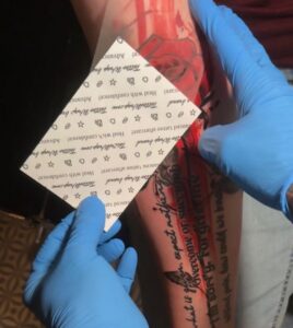 Gloved hands hold a stencil sheet against a tattooed forearm during placement.