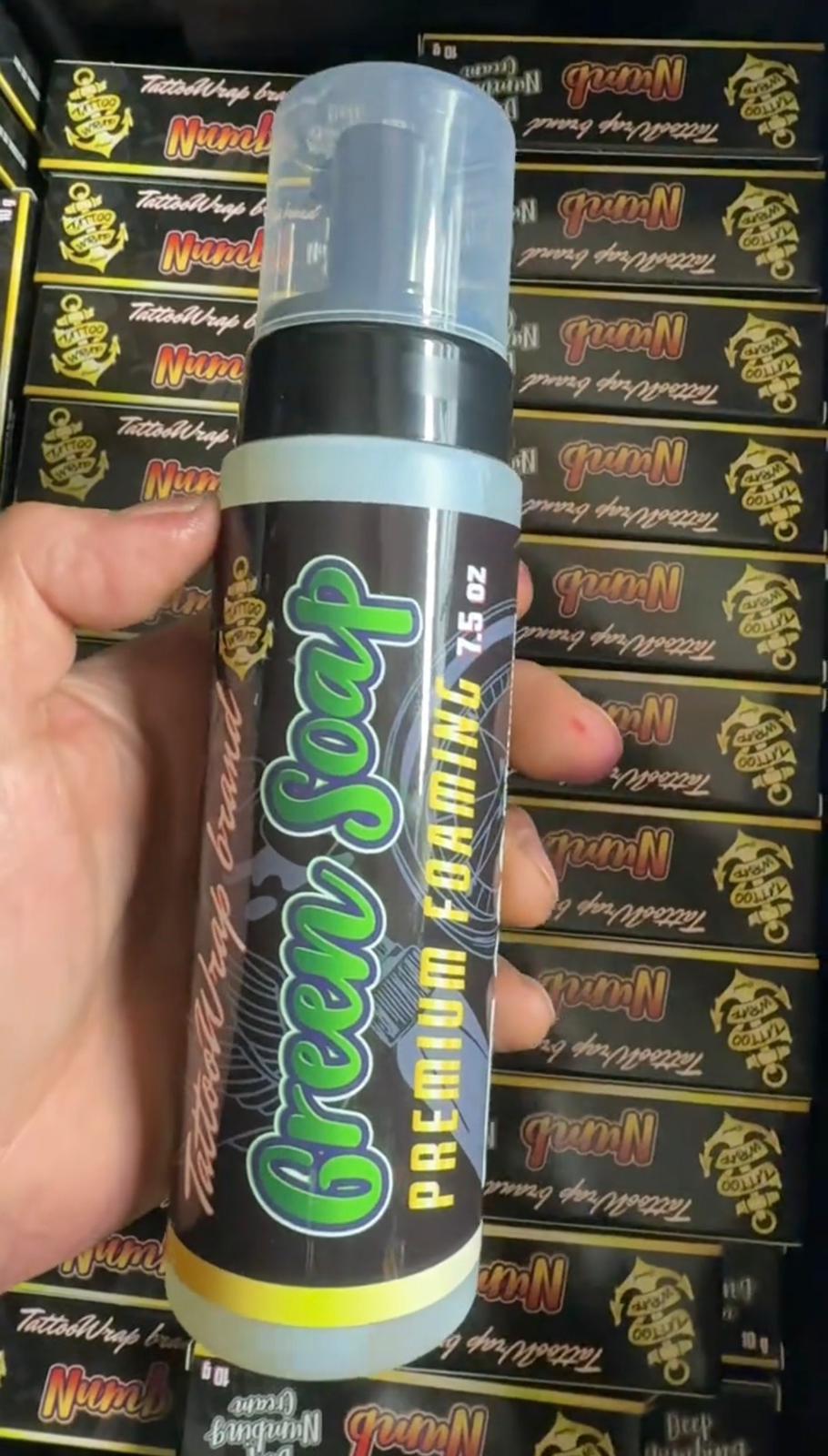Hand holding a tall spray bottle with a clear plastic cap; black label shows bright green script text and yellow vertical lettering, against shelves of boxed products in the background (retail display).