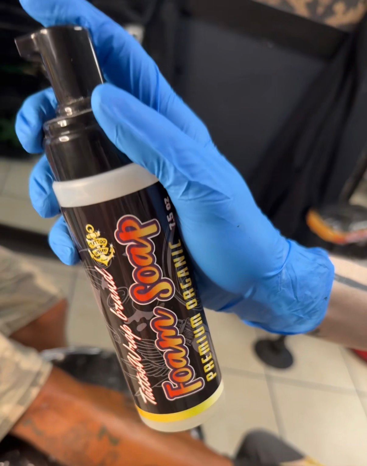 Gloved hand holding a dark spray bottle with a white ring, label shows bold red‑orange text and a yellow cap at the bottom; the person wears blue nitrile gloves.
